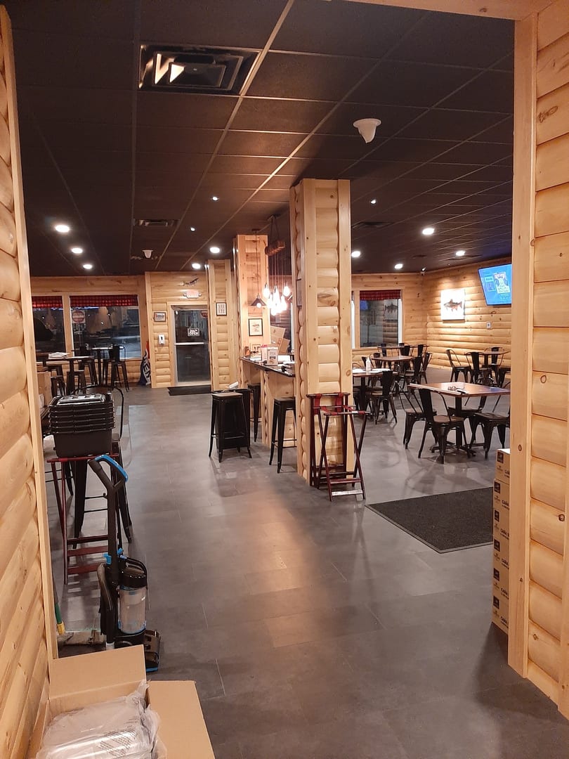 Interior view of a restaurant with wooden walls, dark tables, chairs, and a bar area.