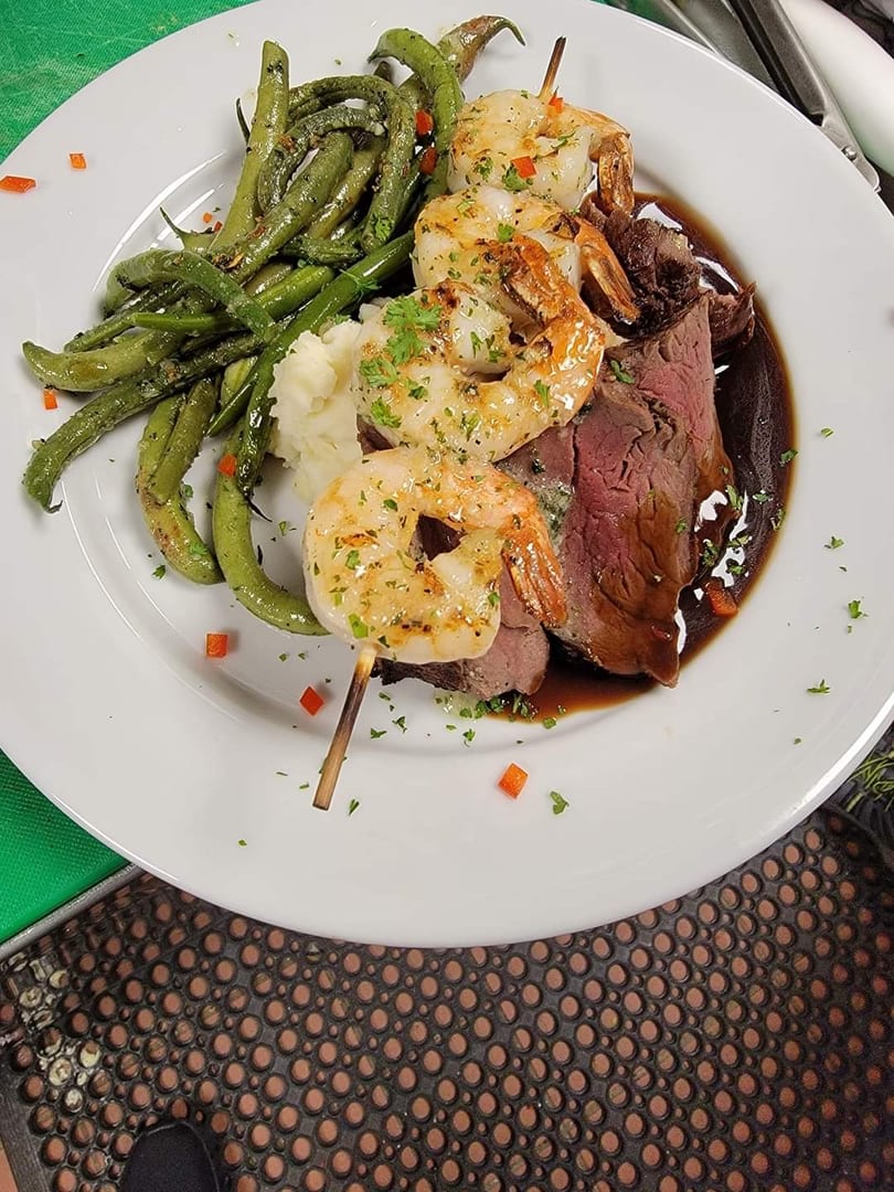 Grilled shrimp skewer, sliced steak with gravy, mashed potatoes, and green beans on a white plate.
