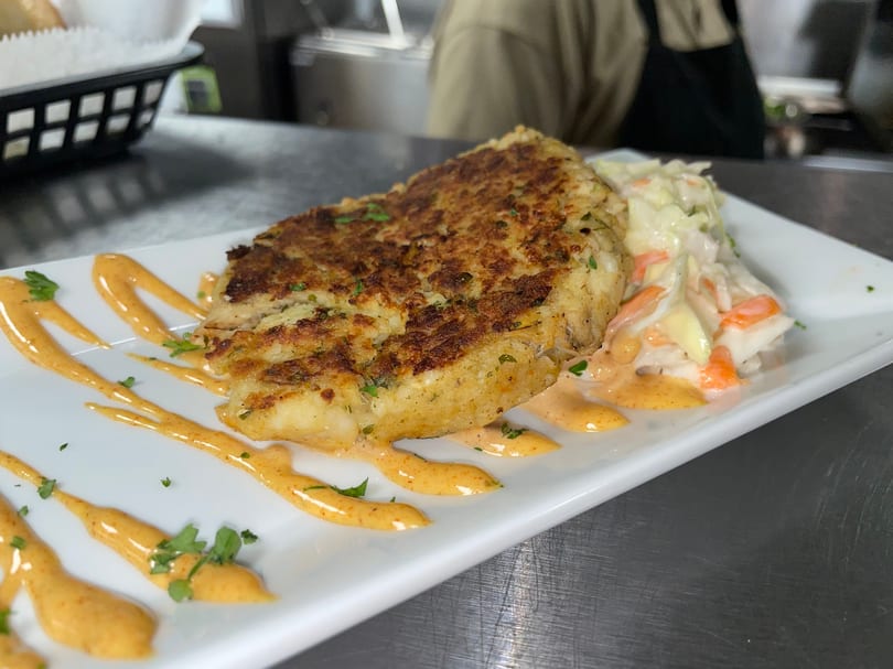 Golden-brown crab cake with coleslaw and a drizzle of orange sauce on a white rectangular plate.