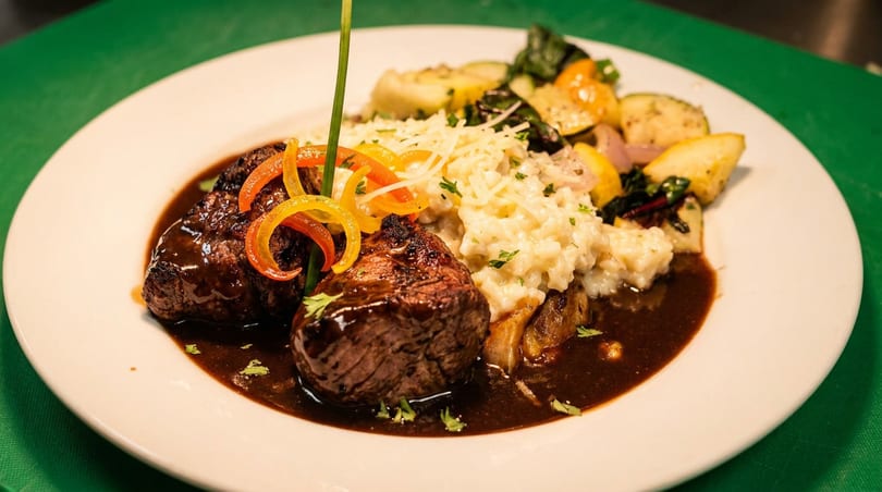 Two cuts of steak with gravy, risotto, and roasted vegetables on a white plate.