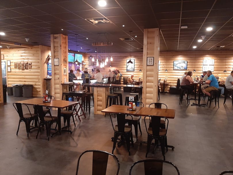 A rustic restaurant interior with wooden walls, dark tables, and patrons dining and socializing.
