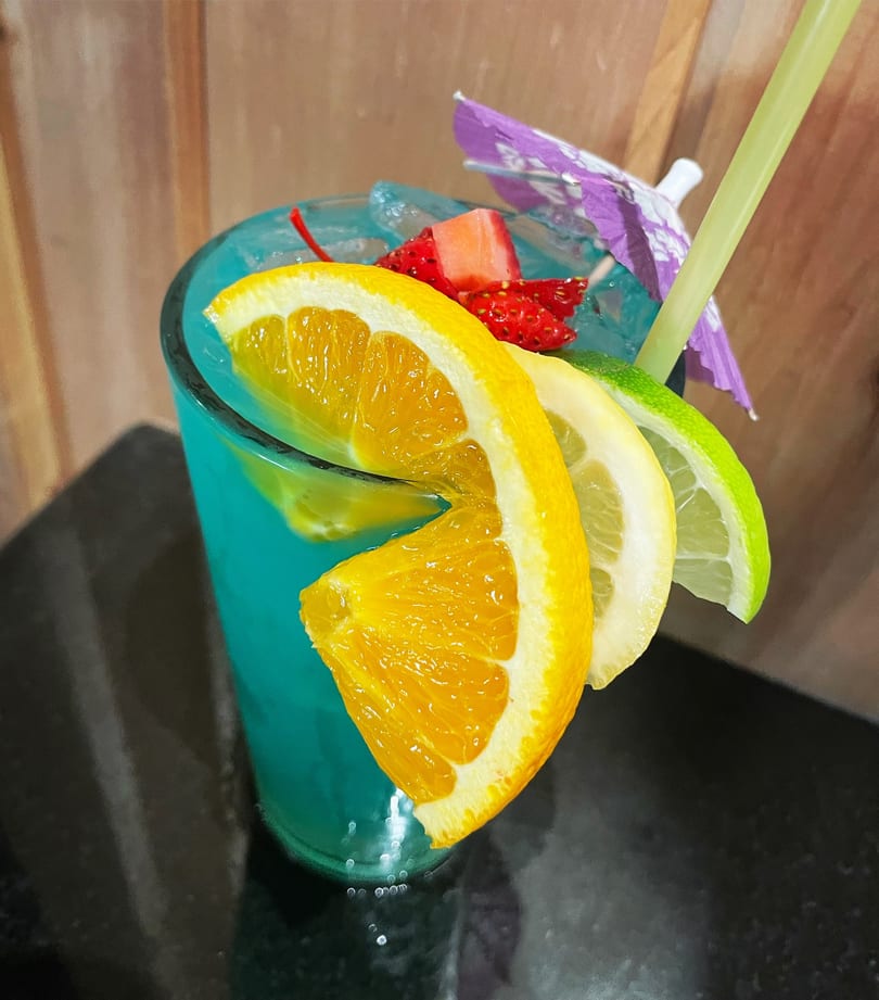 Blue cocktail garnished with orange, lemon, lime slices, strawberries, a cherry, and a paper umbrella.